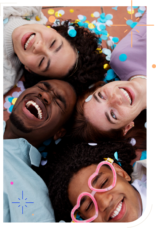 Na fotografia há 2 homens e 2 mulheres enquadrados do ombro para cima, deitadas em círculo, tocando-se pelo topo de suas cabeças. Eles sorriem descontraídos.Um dos rapazes usa um óculos rosa, sem lentes e com armação em formato de corações. Confetes azuis, brancos e amarelos caem sobre eles e se espalham pelo chão.